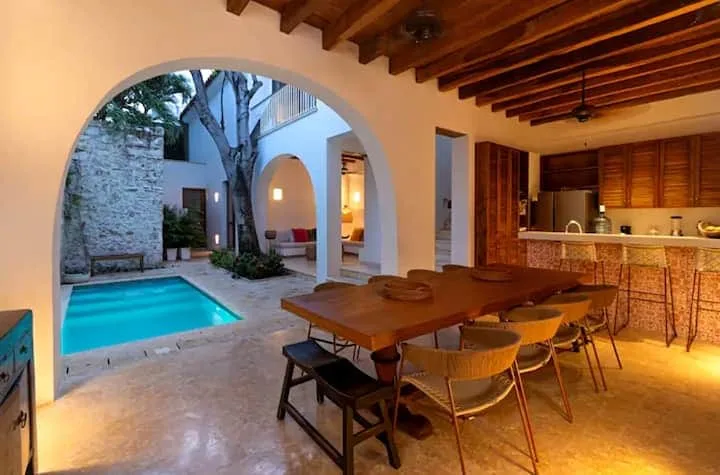 Un comedor abierto da a una piscina en el patio, con una mesa larga bajo vigas de madera.