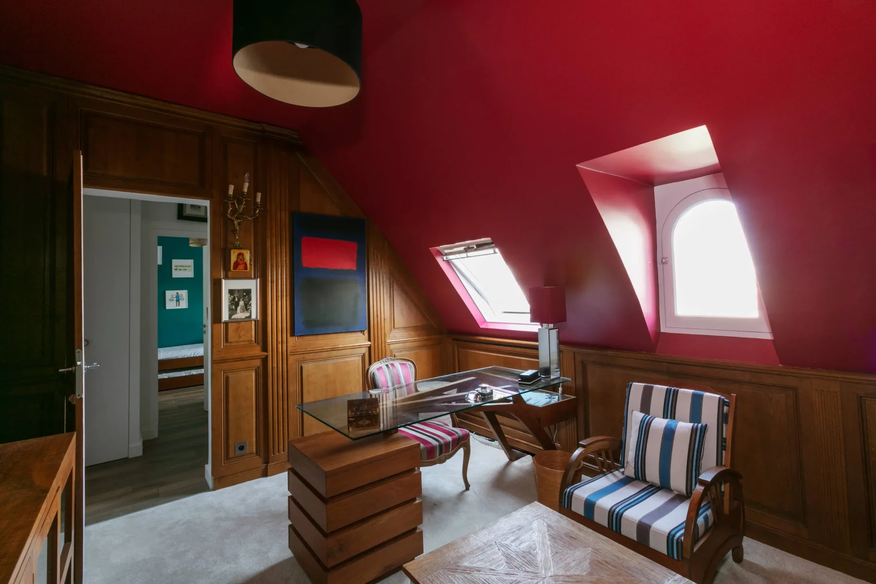 Una habitación abuhardillada de paredes rojas con escritorio y asientos a rayas.