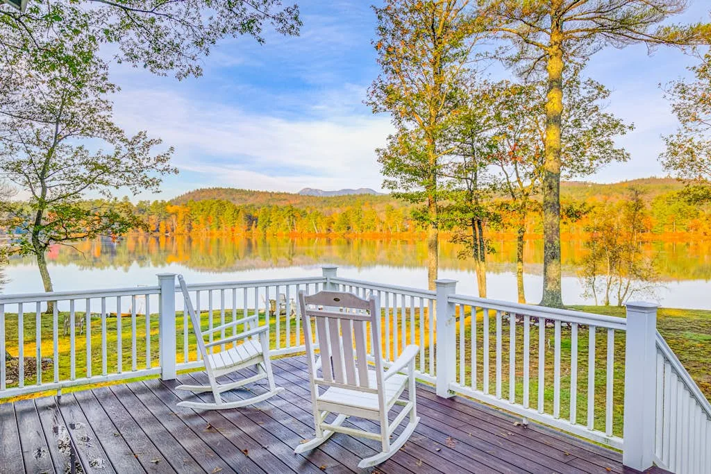 A lakeside deck with white railings and rocking chairs overlooks autumn trees.