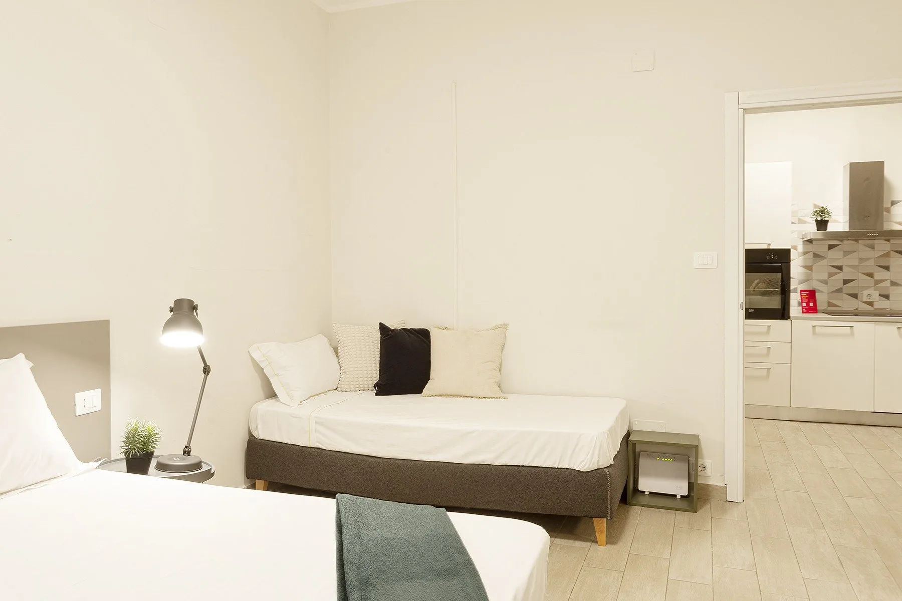 A bedroom with two beds, a bedside lamp, and a view into the kitchen.