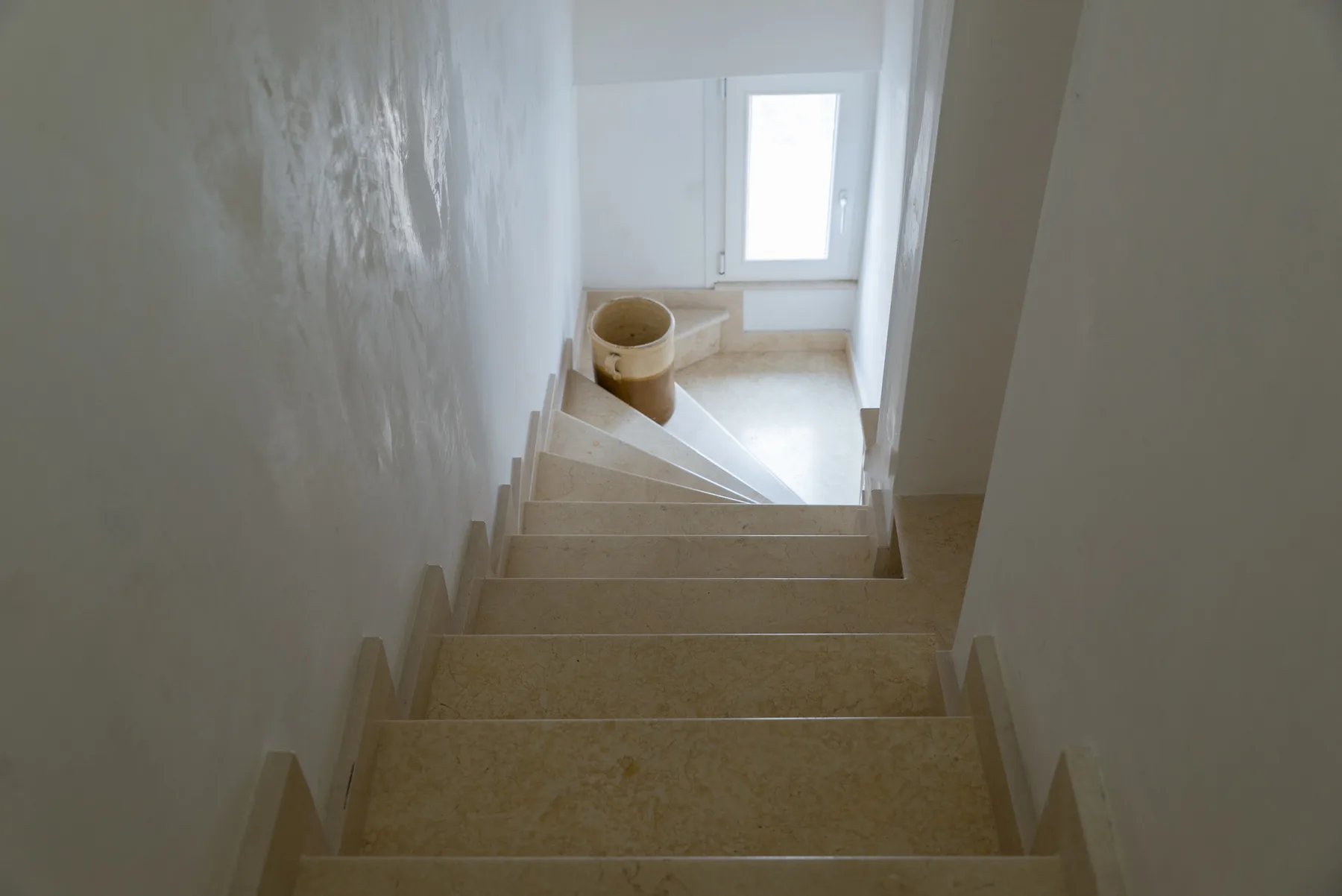 Una escalera estrecha desciende hacia un rellano luminoso con una cesta pequeña y redonda.
