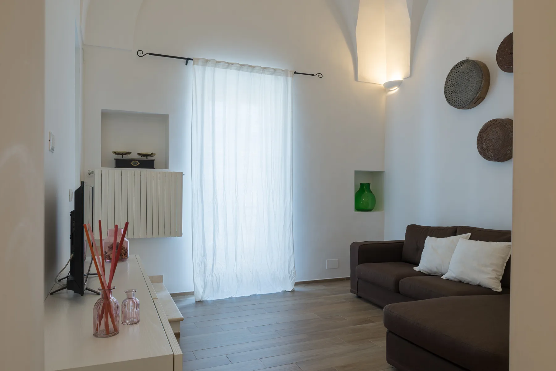 Una sala luminosa con asientos oscuros, un televisor y una ventana con cortinas translúcidas.