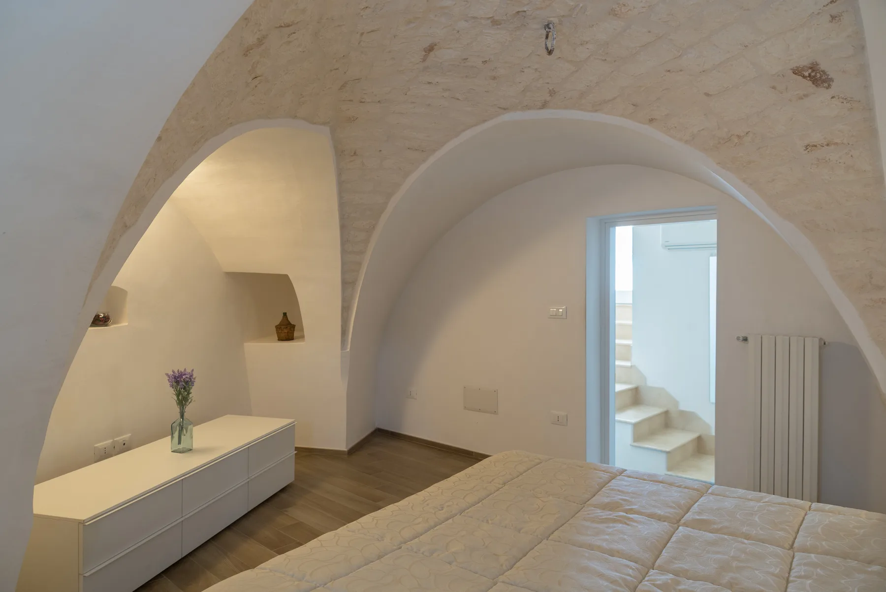 Un dormitorio con un nicho arqueado, un mueble bajo y una puerta hacia una escalera.