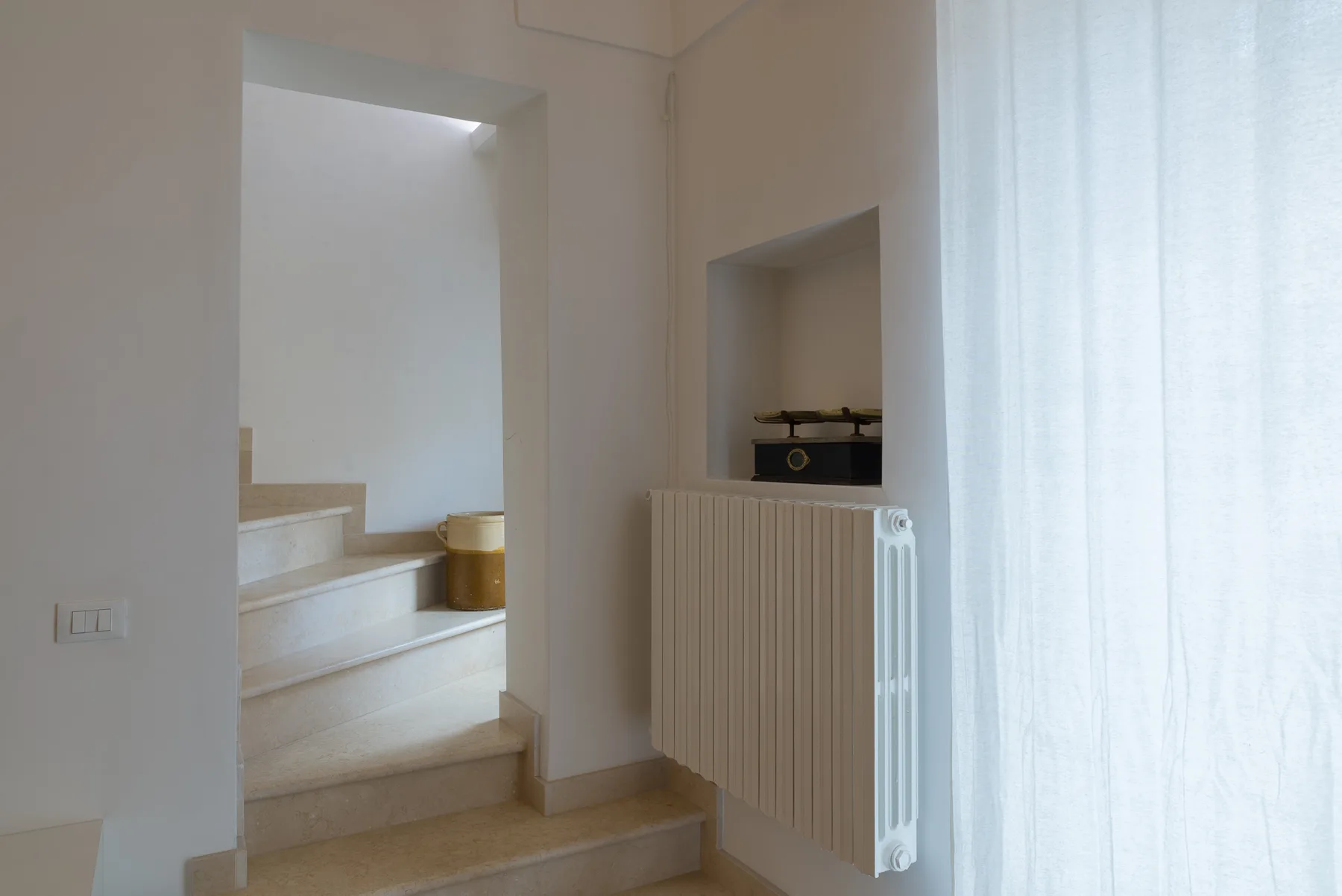 Un pasillo interior luminoso con una escalera y un radiador junto a una cortina translúcida.