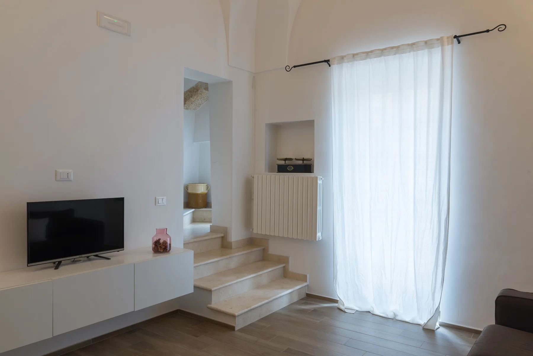 Una sala luminosa con televisor, escalones elevados y cortinas translúcidas.