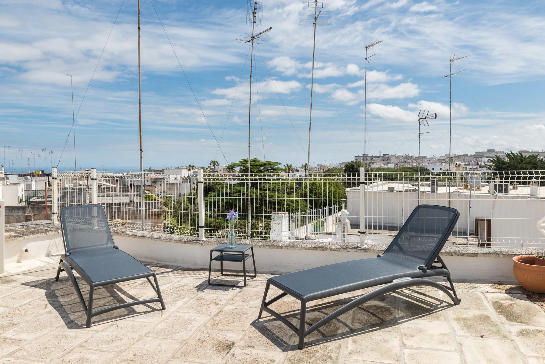 Una terraza en la azotea con tumbonas reclinables, una mesa pequeña y edificios urbanos a lo lejos.