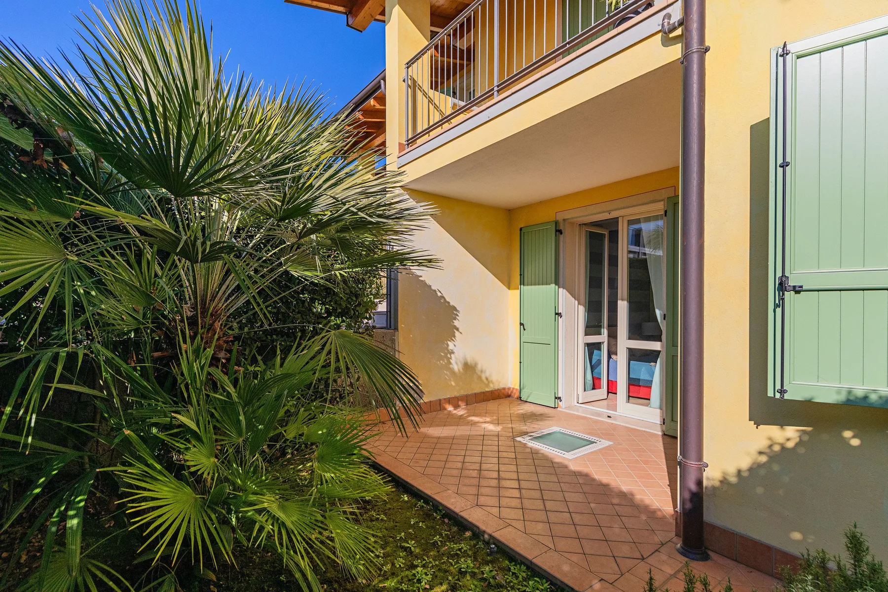 A sunny patio beside a yellow building, with green shutters and leafy plants.