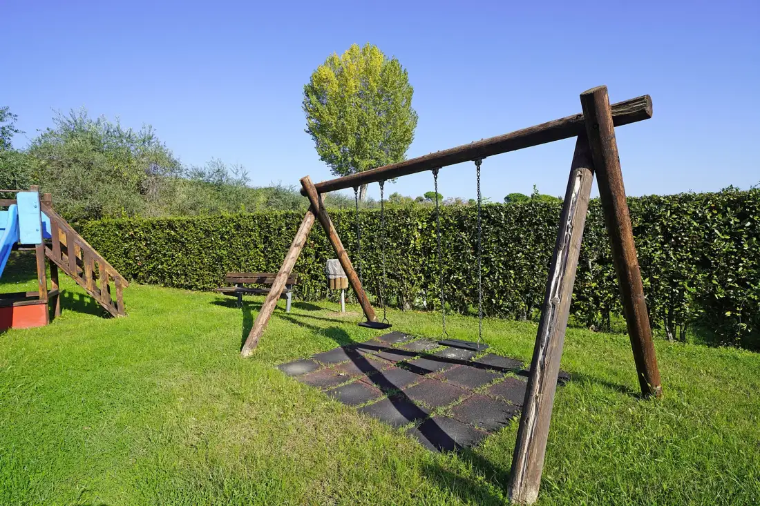 A grassy outdoor play area has a wooden swing set beside a hedge.