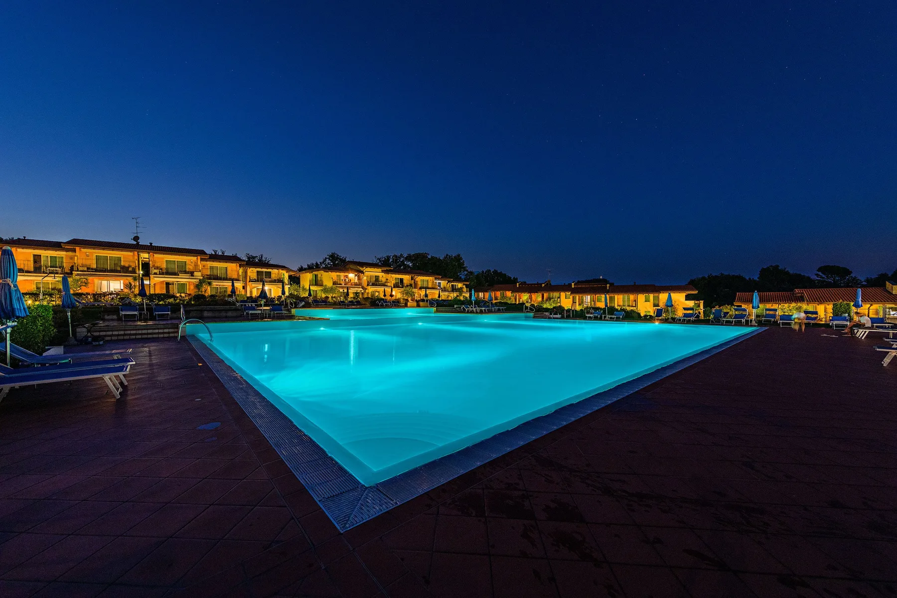 An outdoor swimming pool glows at dusk beside a deck and illuminated buildings.