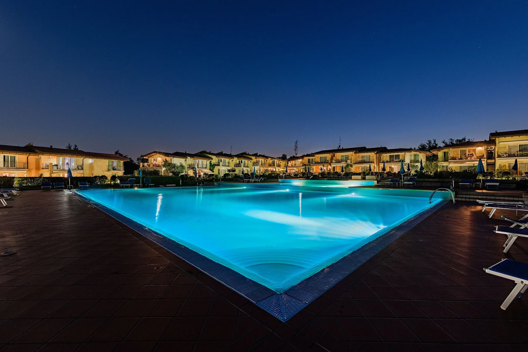 A lit swimming pool sits among low buildings beneath the evening sky.