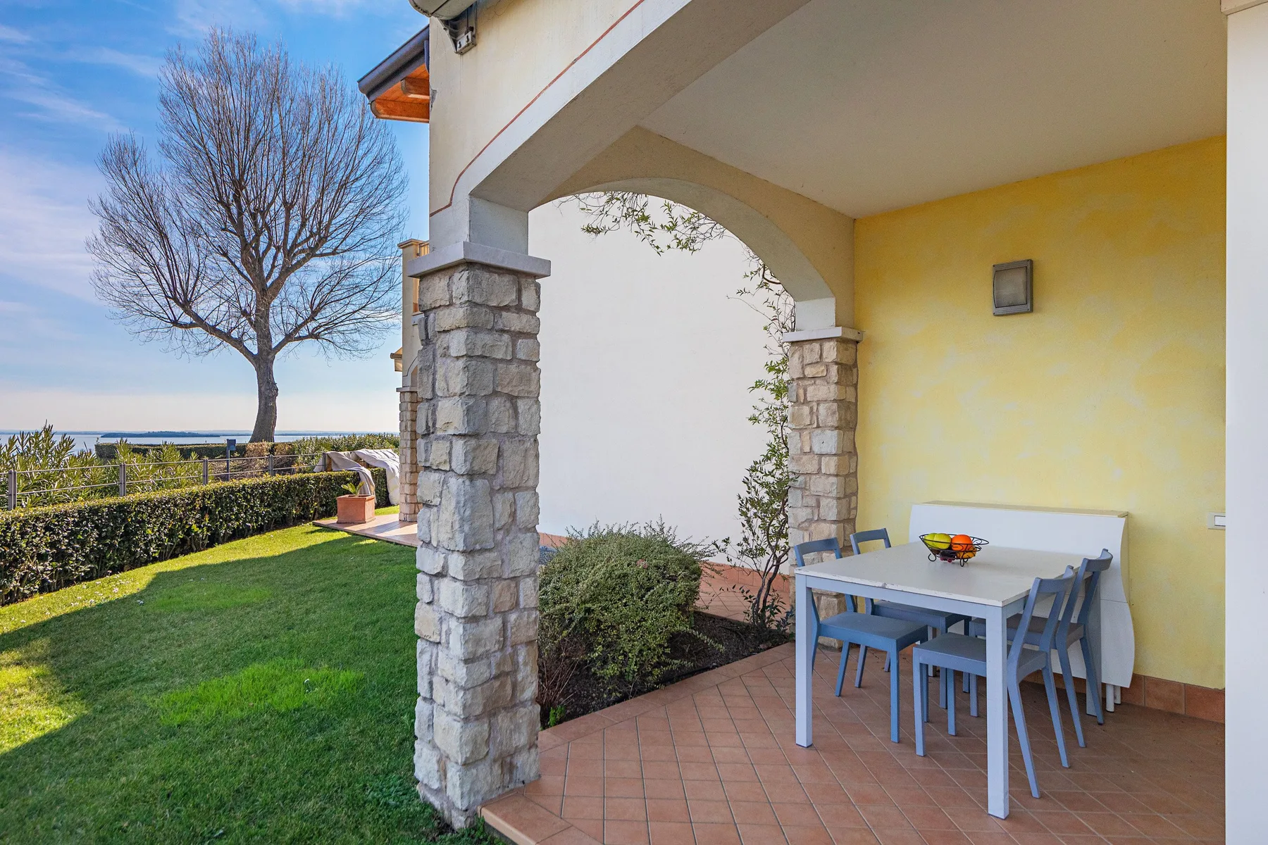 A covered patio with outdoor seating overlooks a green lawn and leafless tree.