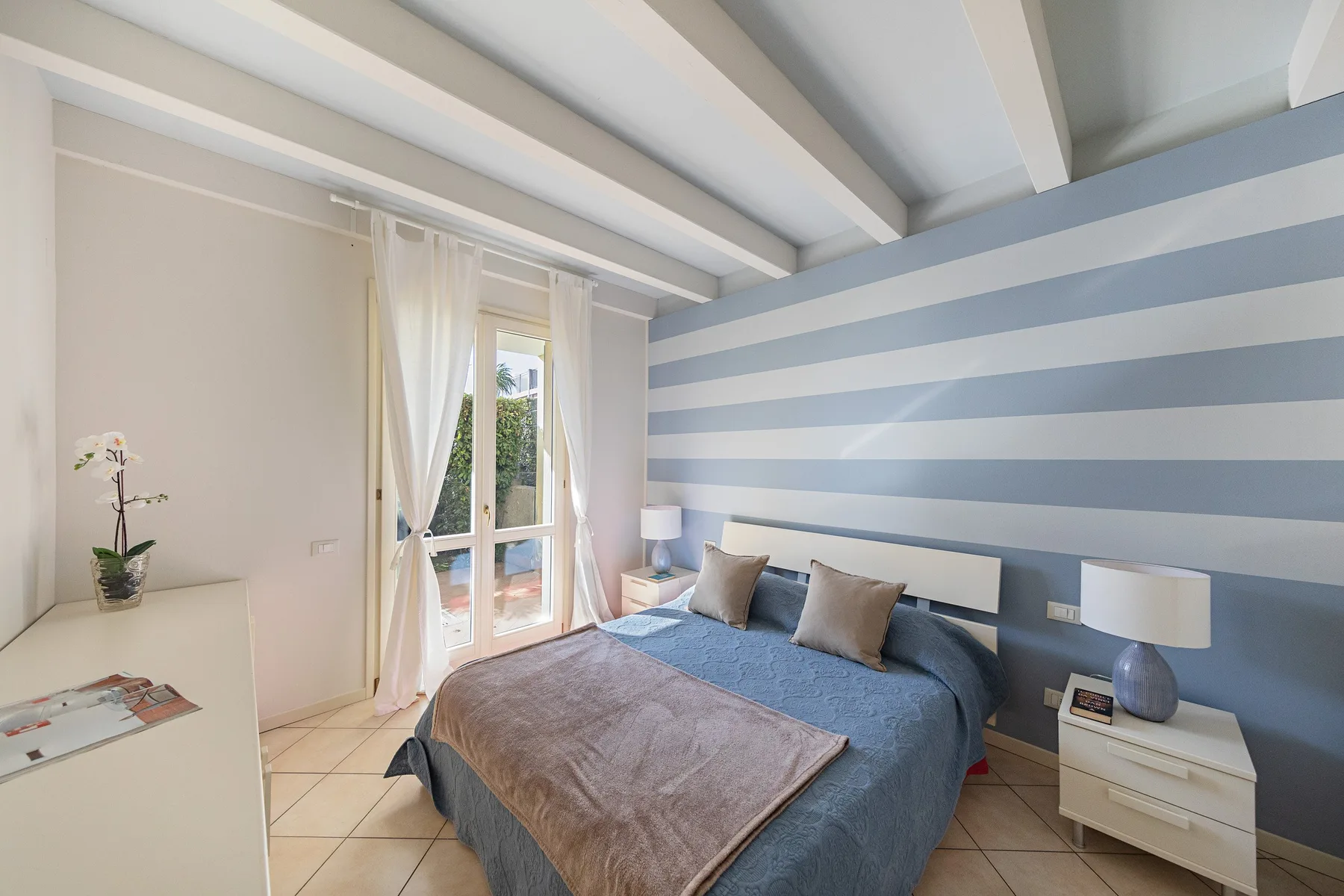 A bedroom with a blue bed and striped walls beside a curtained window.