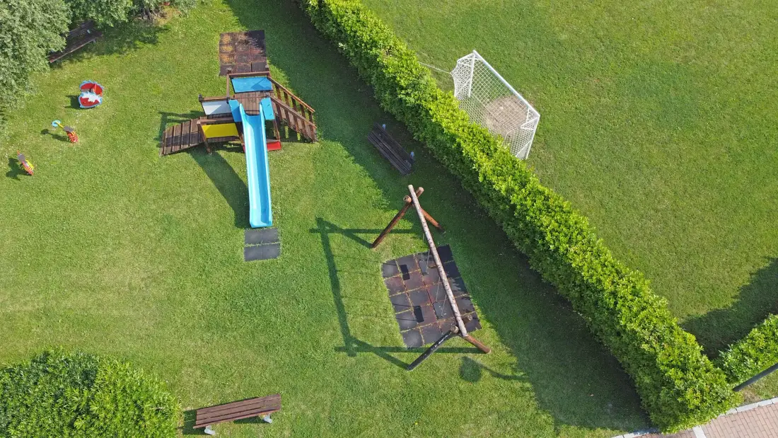 A grassy play area has a wooden slide, swings, and a small goal.