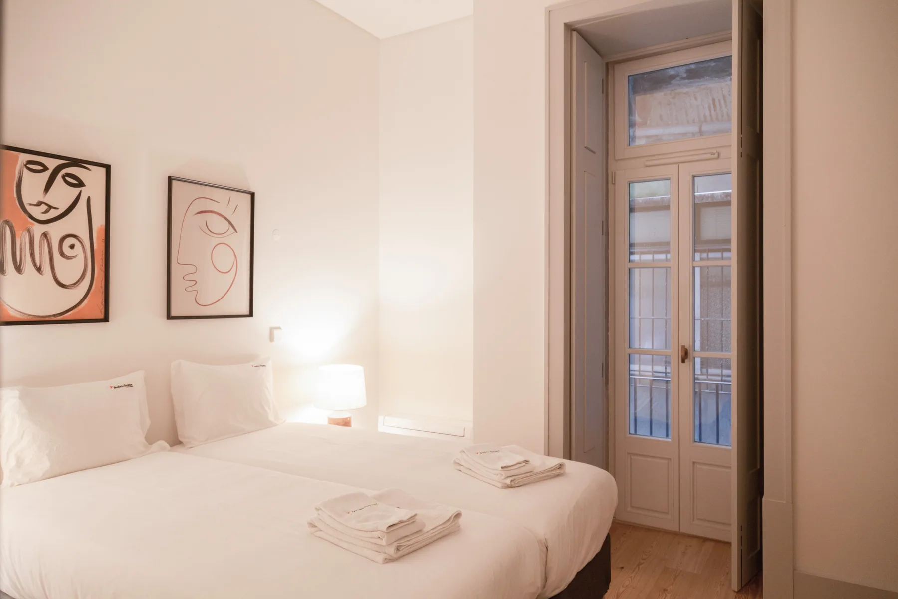 A bright bedroom with white bedding, framed artwork, and a tall glazed door.