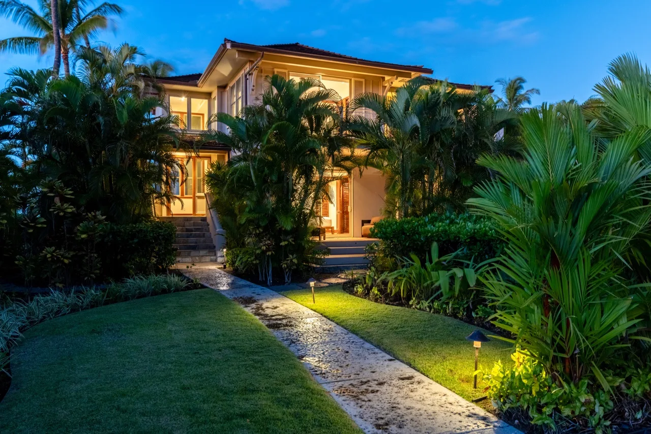 A warmly lit house stands behind tropical landscaping, with a path leading to the entrance.