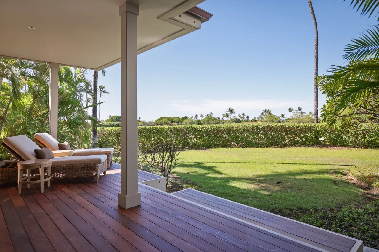 A covered outdoor deck with cushioned seating overlooks a lawn and hedge.