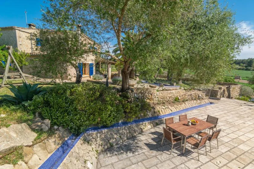 A tiled outdoor patio with a dining table sits beside trees and a stone house.