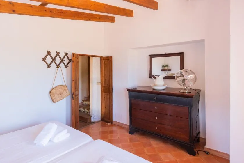A bedroom with a dark dresser, a standing fan, and exposed wooden ceiling beams.