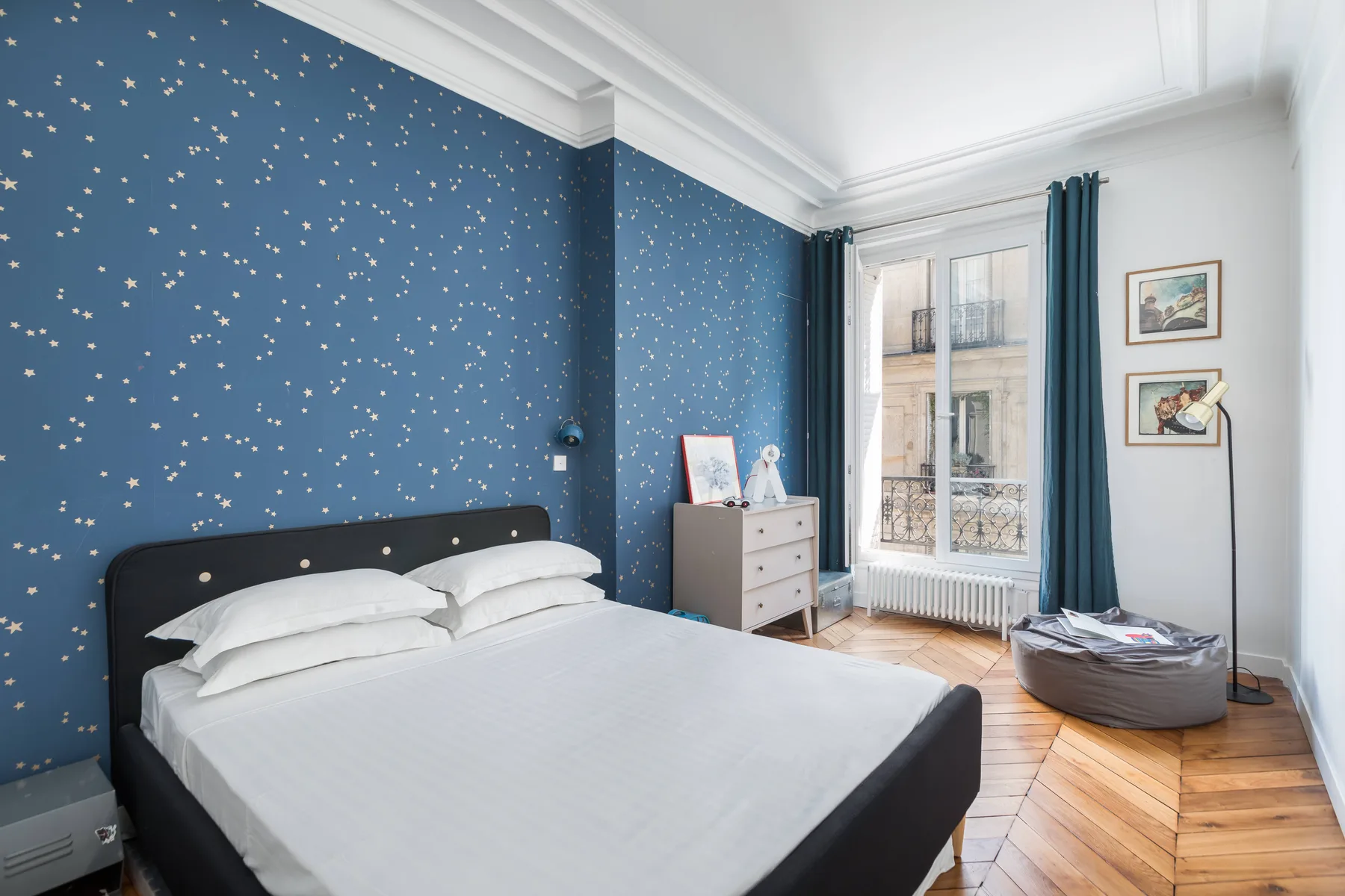 Un dormitorio con una cama, paredes azules estampadas y una ventana alta.