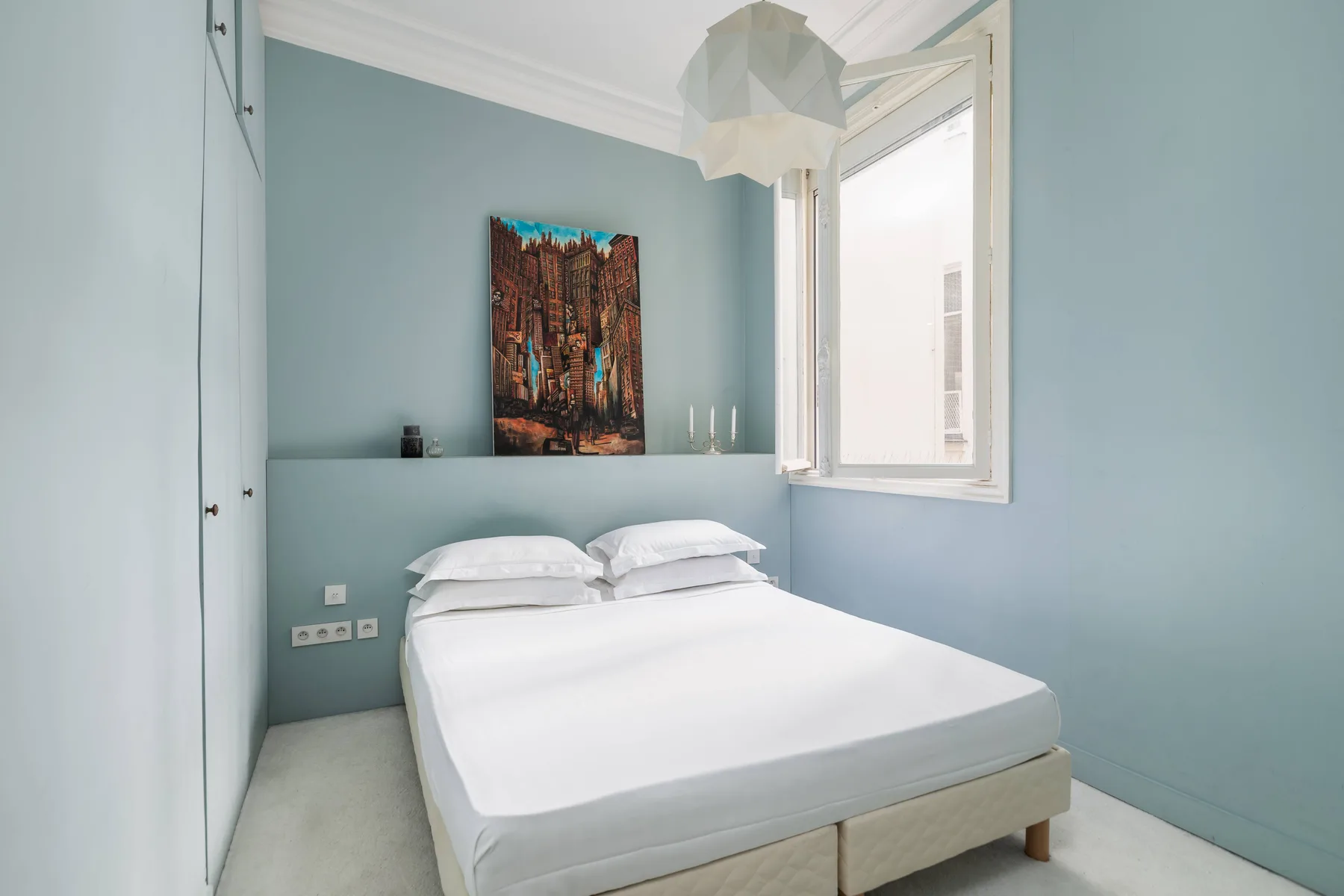 A bedroom with a neatly made bed, pale blue walls, and a bright window.