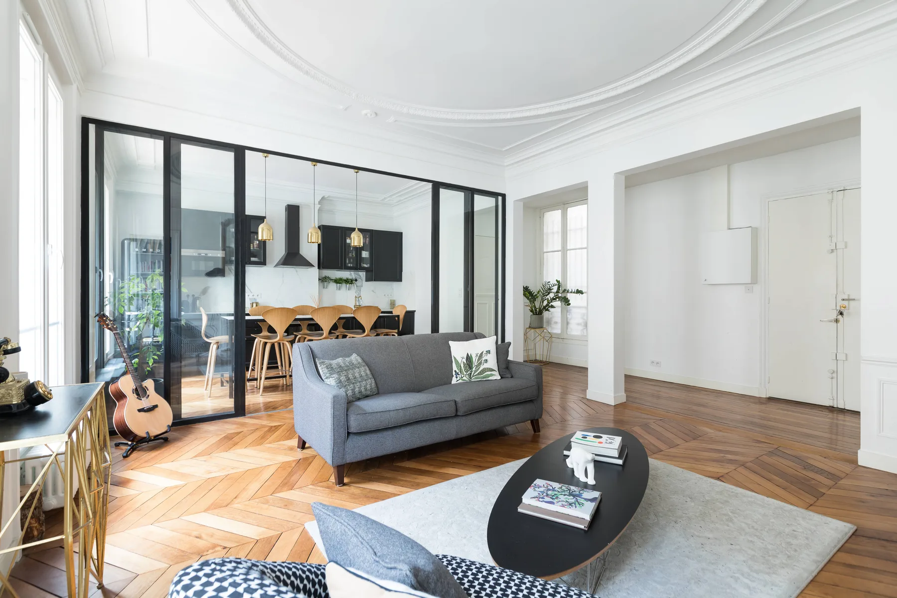 Una sala luminosa con un sofá gris y una mampara de vidrio que separa el comedor.