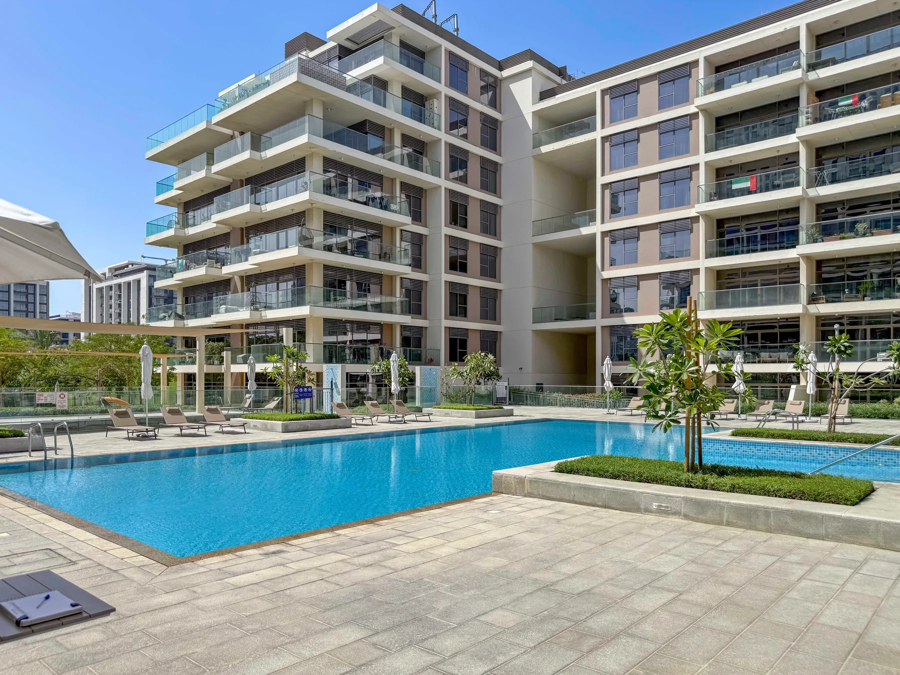 Una piscina está junto a edificios de apartamentos modernos y zonas pavimentadas al aire libre.
