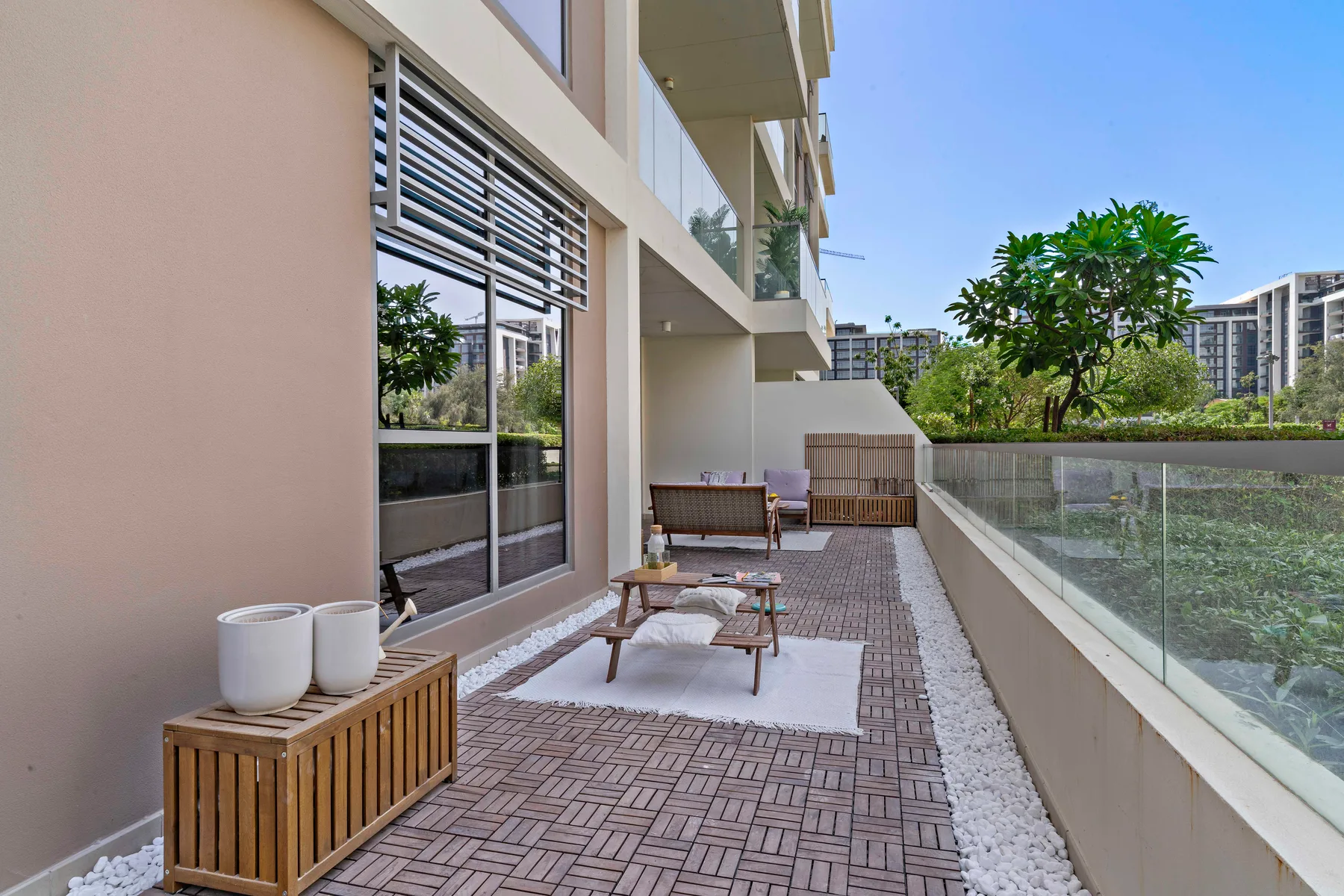 Una terraza exterior embaldosada con asientos y una barandilla de vidrio da a jardines.
