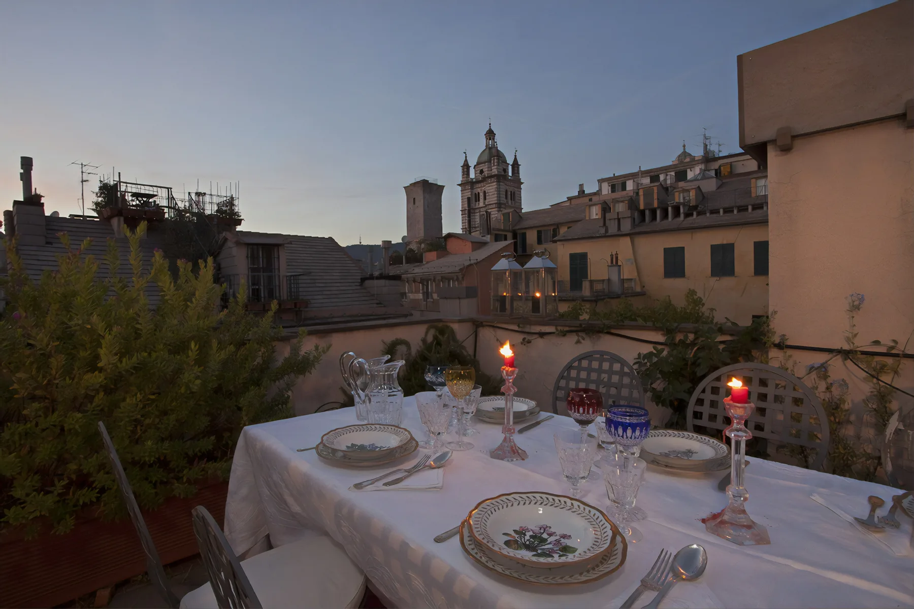A rooftop dining table is set with plates and lit candles, overlooking nearby buildings.