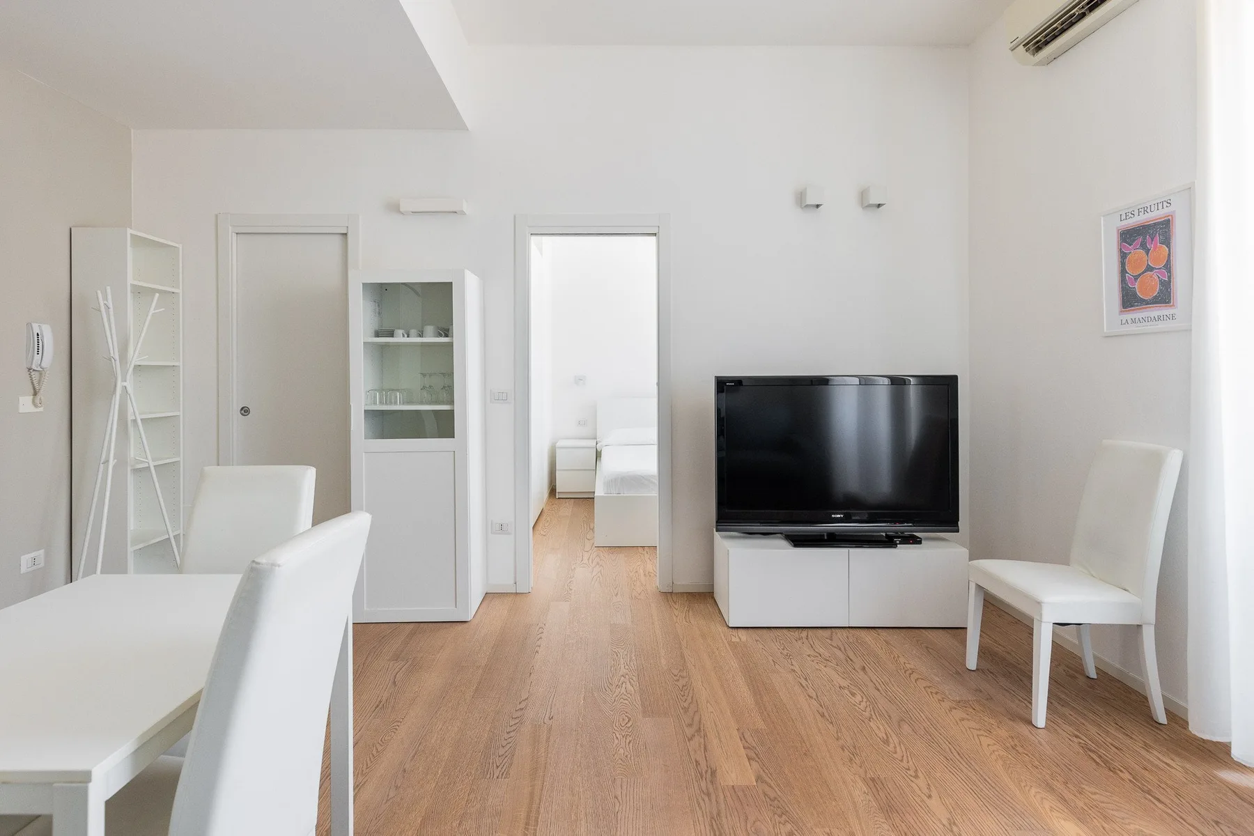 Una luminosa sala de estar con televisor, asientos blancos y una puerta hacia un dormitorio.