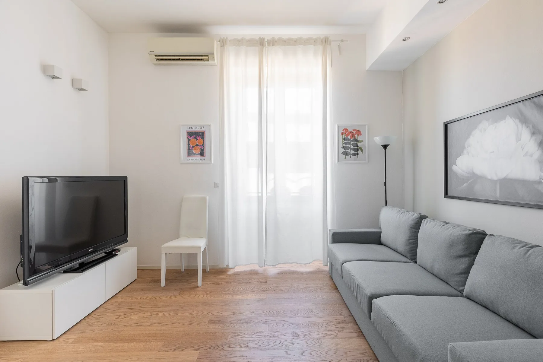 Una sala luminosa con un sofá gris y un televisor junto a una ventana con cortinas.