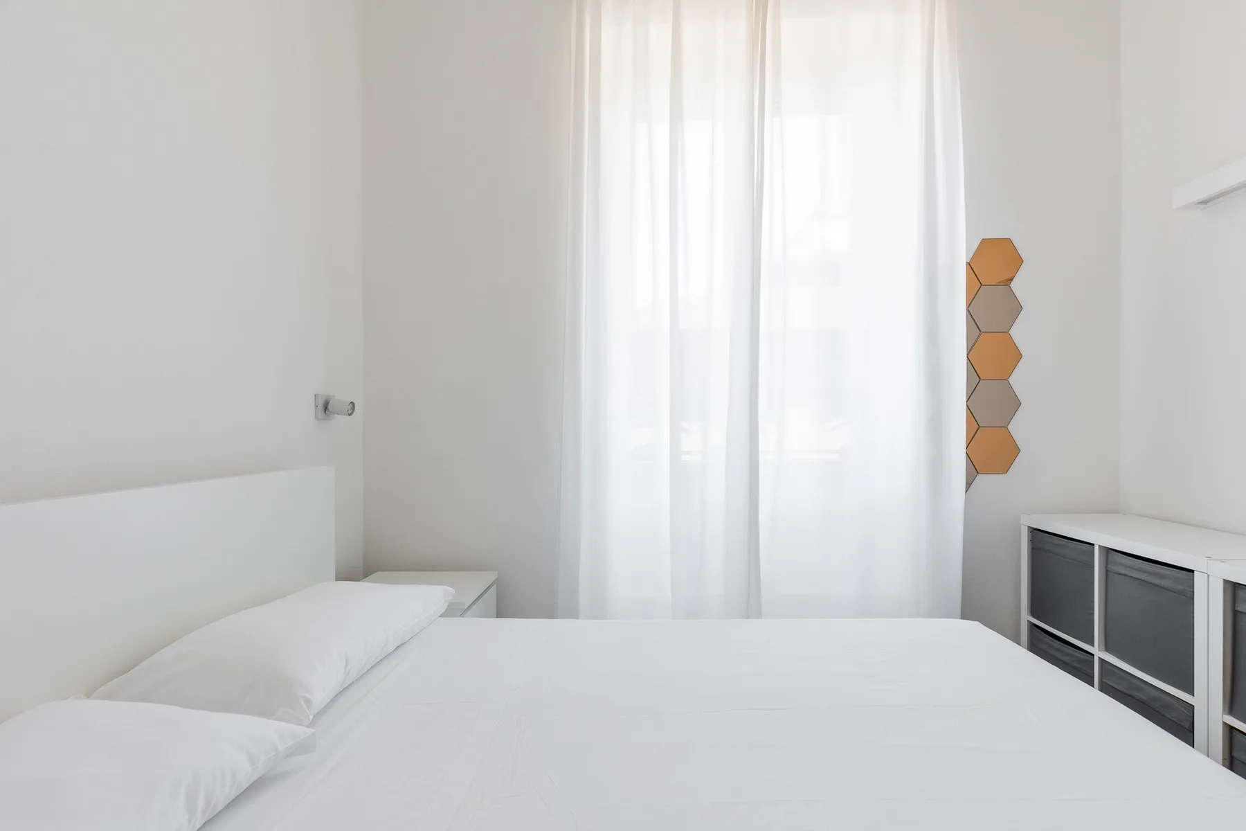 Un dormitorio luminoso con una cama blanca bajo una ventana alta con cortinas.