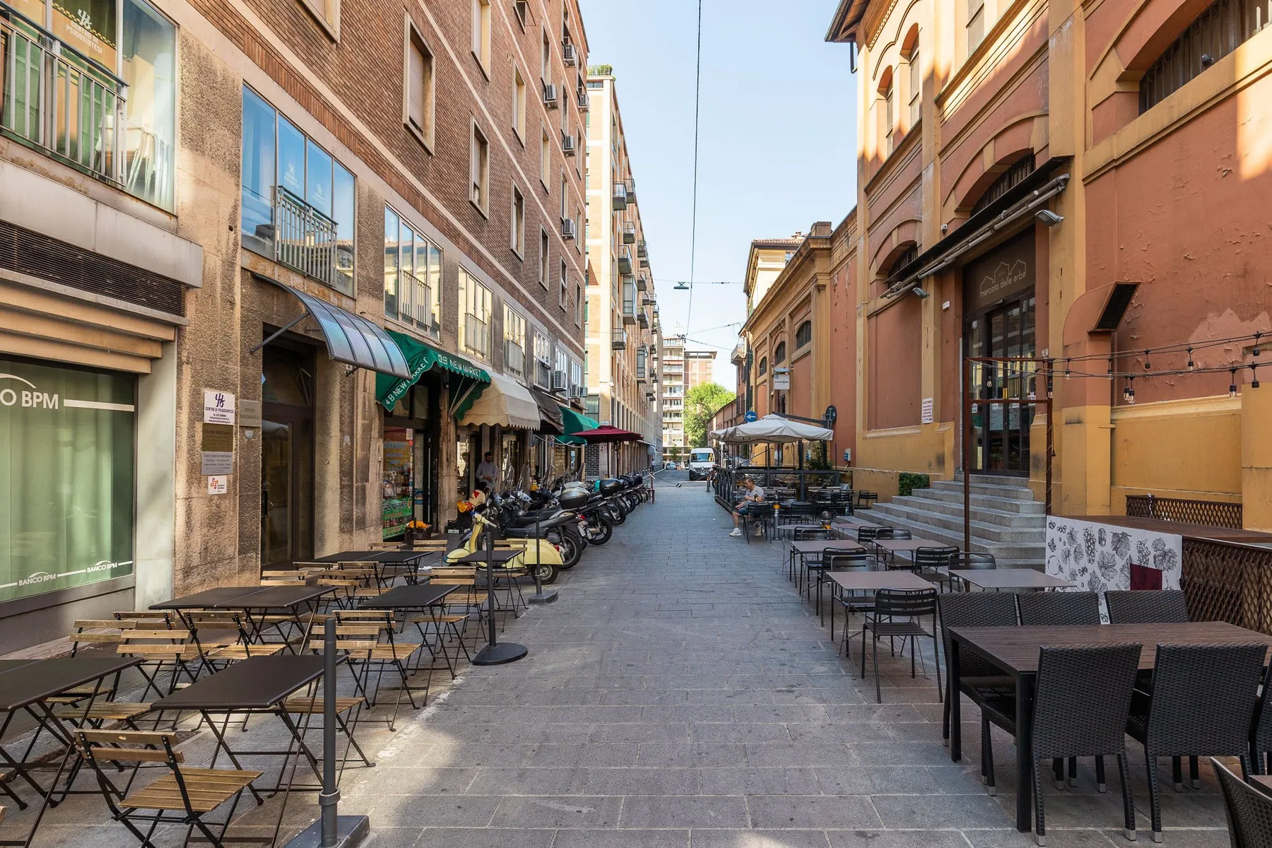 Una calle peatonal estrecha está flanqueada por mesas de café al aire libre entre edificios altos.