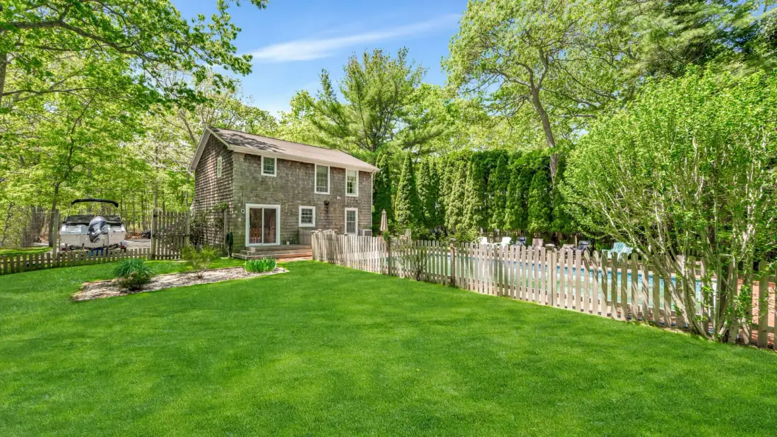 A two-story house stands beside a fenced lawn, surrounded by leafy trees.