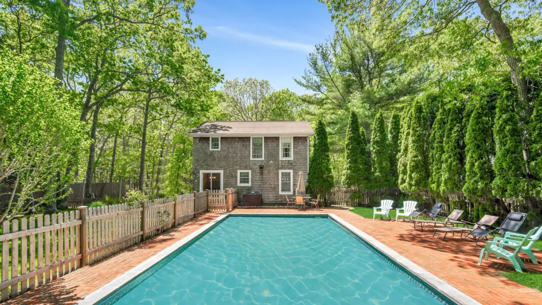 Outdoor swimming pool beside a house, surrounded by trees and outdoor seating.