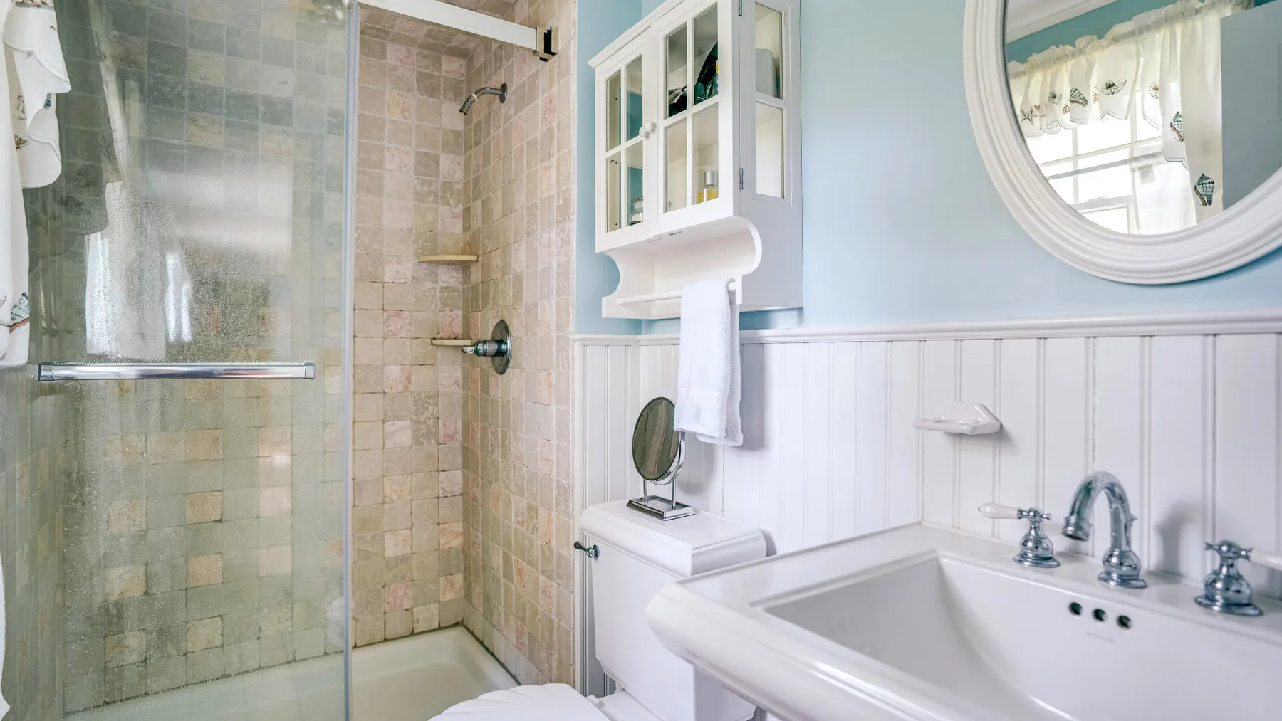 A bathroom with a glass shower, a sink, and a round wall mirror.