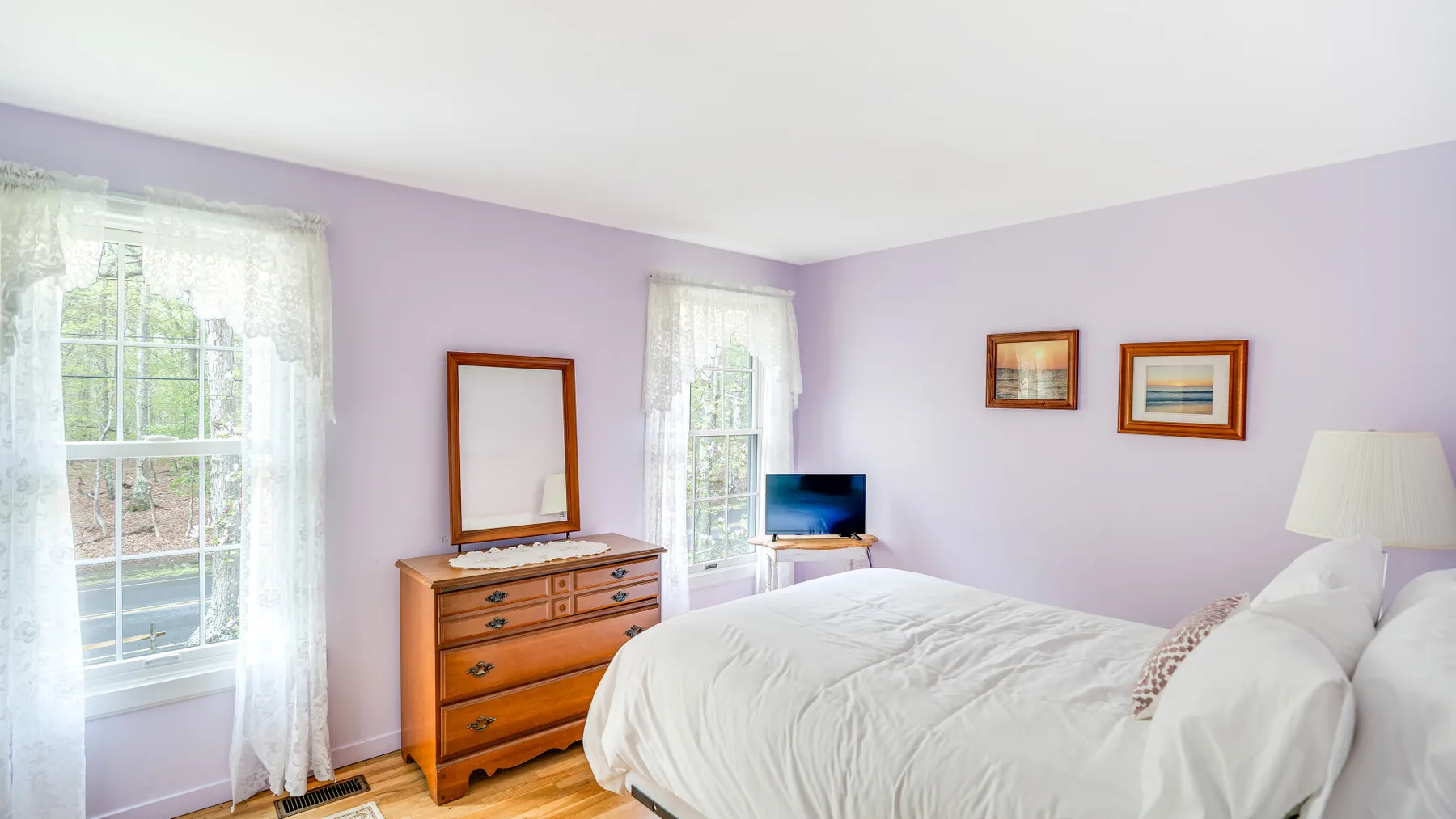 A bedroom with a white bed, wooden dresser, and lavender walls.