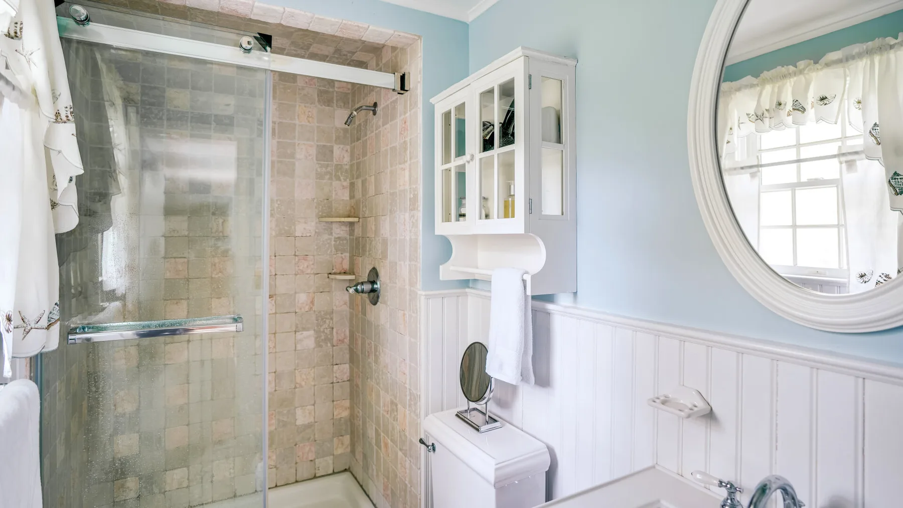 A bathroom with a glass shower and a large round wall mirror.