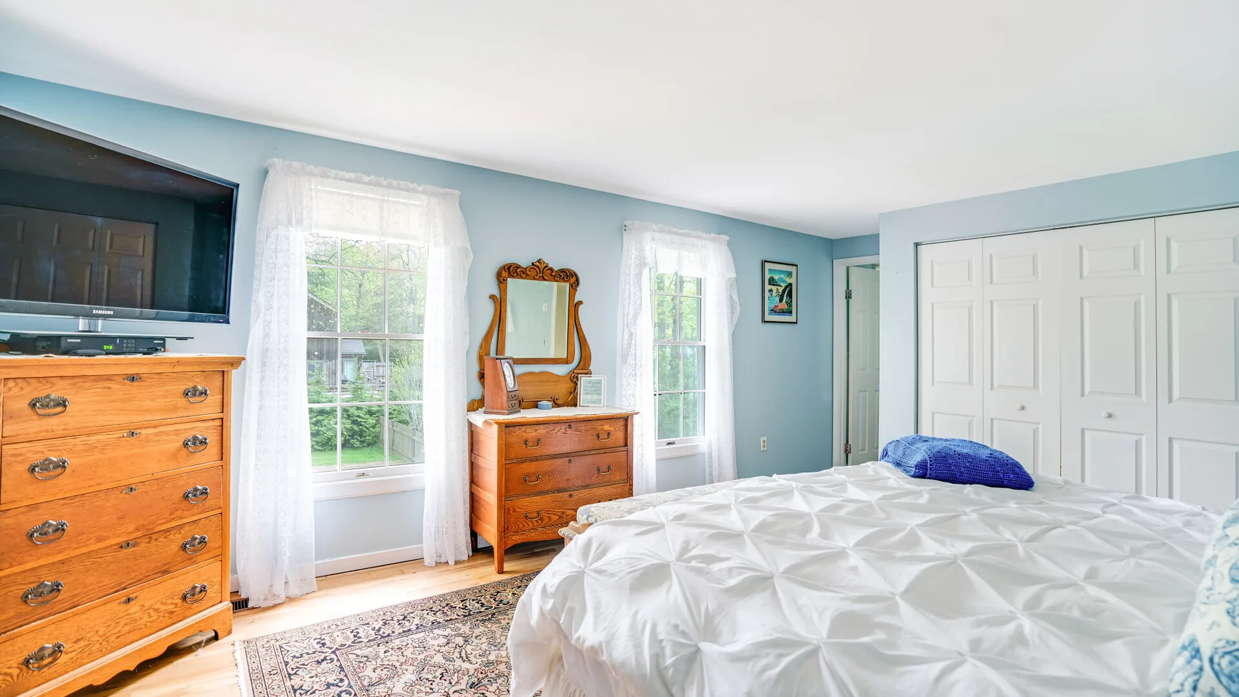 A bedroom with a white bed, wooden dressers, and windows overlooking greenery.