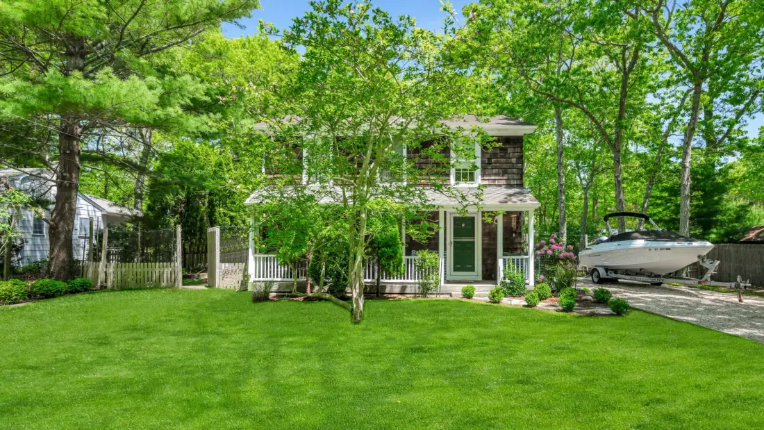 A house with a white porch stands behind a broad green lawn and leafy trees.