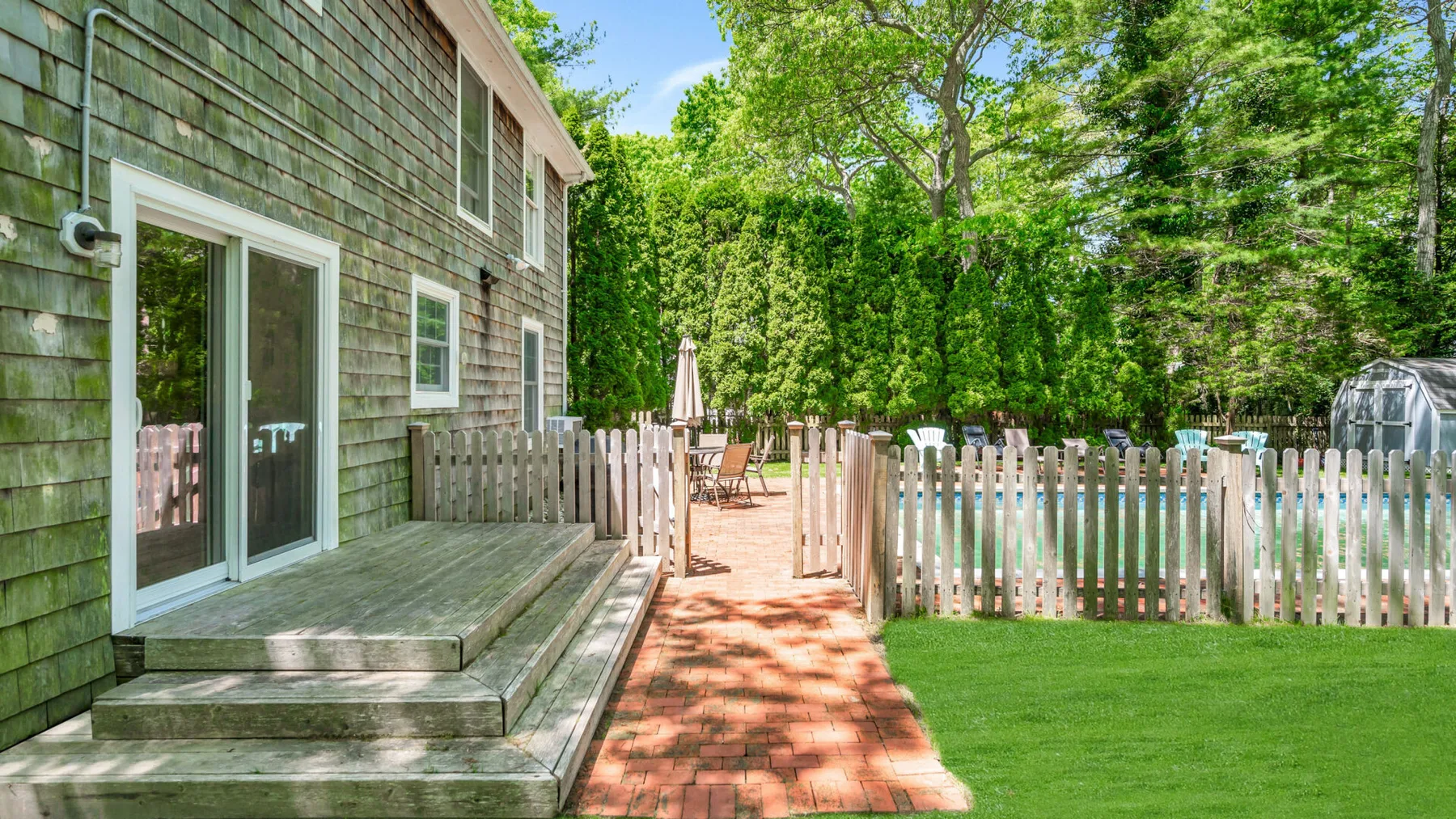 A brick patio beside a house opens onto a fenced green yard surrounded by trees.