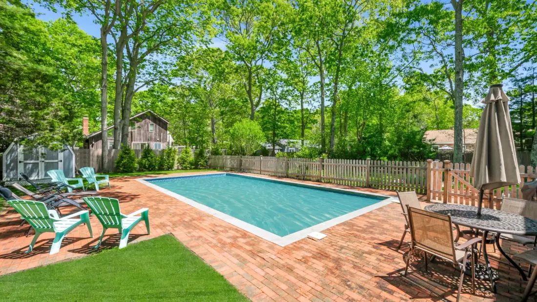 An outdoor swimming pool sits on a brick patio surrounded by trees and seating.