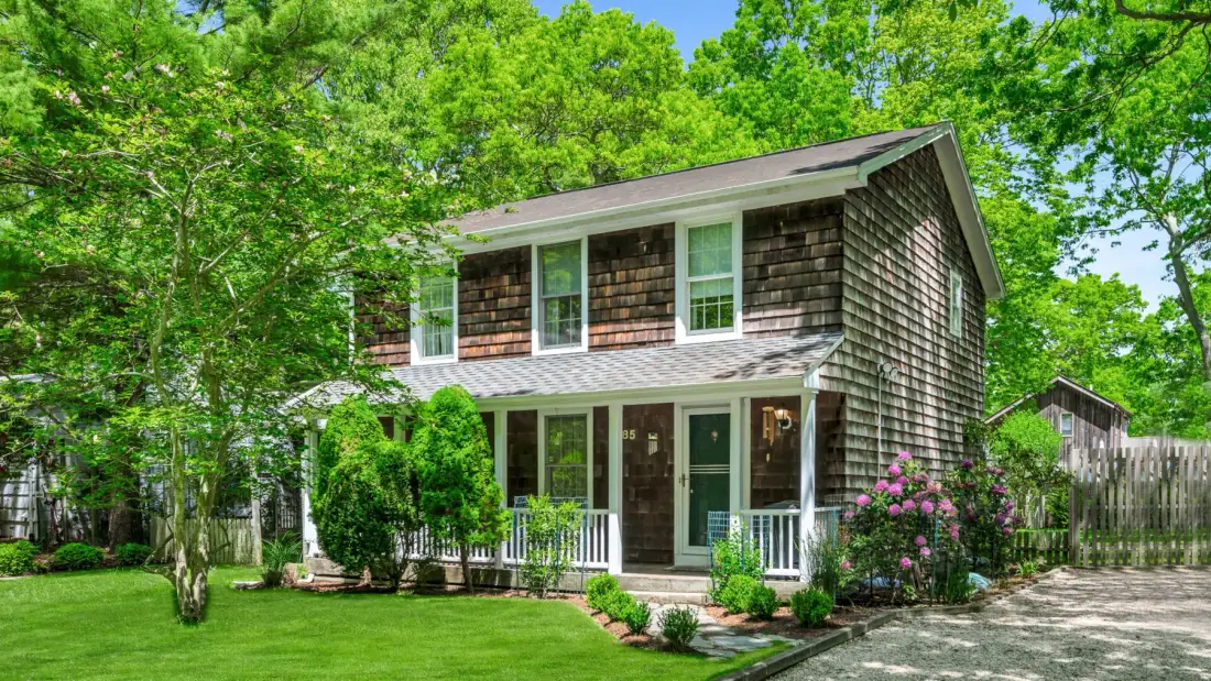 A two-story house stands among trees, with a lawn and flowering shrubs in front.