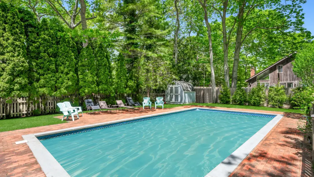 An outdoor swimming pool is surrounded by a brick patio and leafy trees.