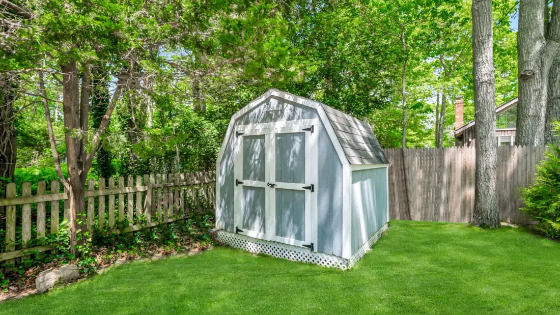 A small garden shed stands on a green lawn among trees and wooden fences.