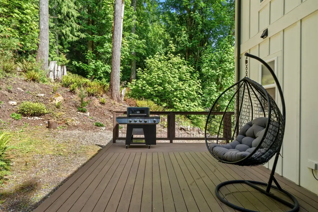 Una terraza de madera da a un jardín arbolado, con una silla colgante acolchada.