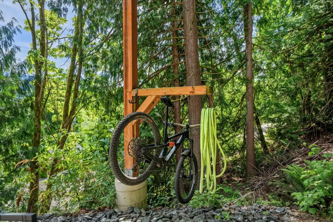 Una bicicleta y una manguera enrollada cuelgan junto a una estructura de madera entre árboles.