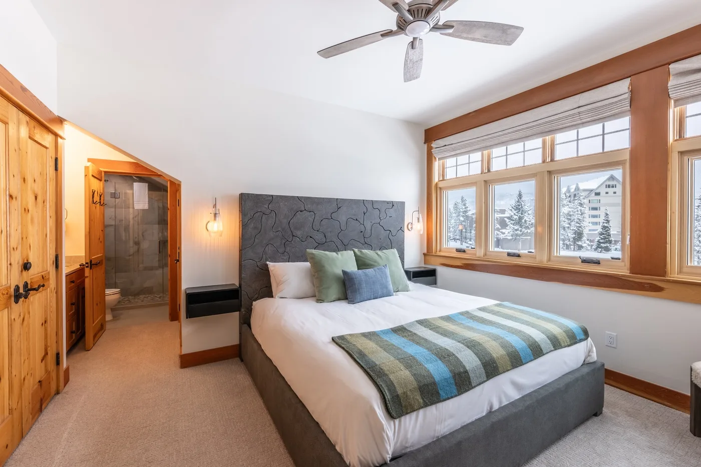 A bedroom with a bed and large windows overlooking trees and nearby buildings.
