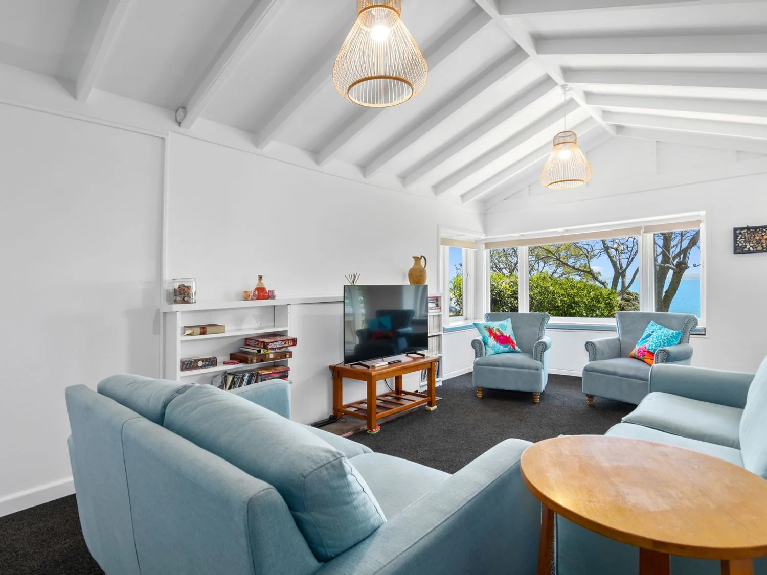 Una sala de estar con asientos azules, un televisor y grandes ventanas con vistas a árboles.