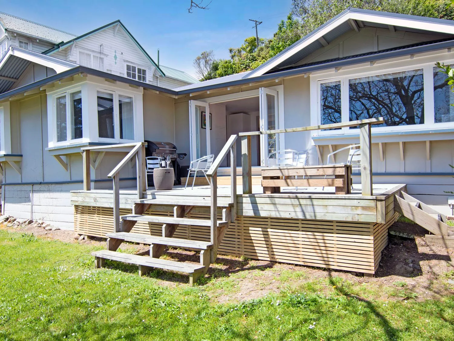 Una terraza exterior de madera adosada a una casa, con escaleras, sillas de metal, una parrilla y una mesa en Bayview Bungalow - Nelson Holiday Home, en Nelson.