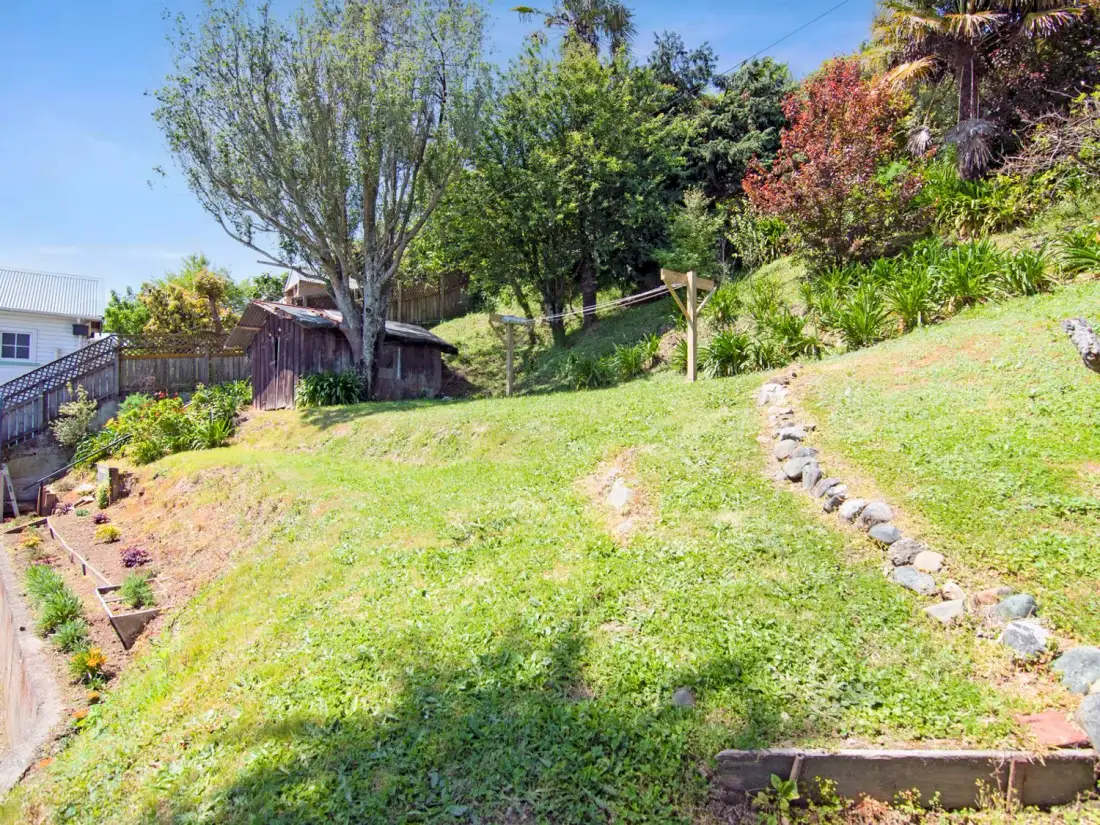 Un jardín trasero con una pendiente cubierta de césped, árboles y un pequeño cobertizo.